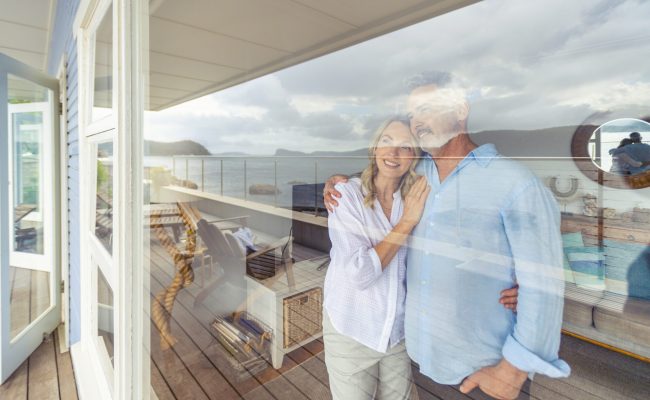 Mature couple looking at the view in their waterfront home. They look happy and contented. They are embracing looking through a window. The ocean can be seen in the reflection